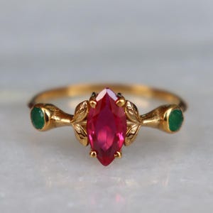 Może przedstawiać: Złoty pierścionek z centralnym rubinem o szlifie markizy, otoczonym dwoma szmaragdami. Pierścionek ma delikatny, liściasty wzór wokół rubinu. Oprawa jest z polerowanego złota. Pierścionek jest umieszczony na marmurowym tle.