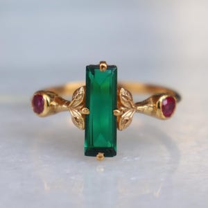 Peut inclure: Une bague en or ornée d'une pierre verte rectangulaire au centre, flanquée de deux petites pierres rouges rondes. La bague présente un motif délicat en forme de feuilles sur la monture. La bague est posée sur un fond clair.