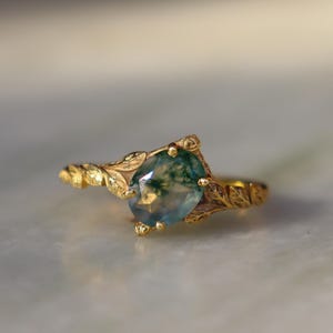 Peut inclure: Une bague en or ornée d'une pierre précieuse ovale verte et bleue. La bague présente des détails complexes en forme de feuilles autour de la pierre et le long de l'anneau. L'arrière-plan est flou.