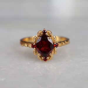 May include: A gold ring featuring a large oval-cut deep red garnet at the center, surrounded by smaller round red garnet accents. The metal band and the setting are crafted with a textured leaf motif that encircles the central gemstone cluster.