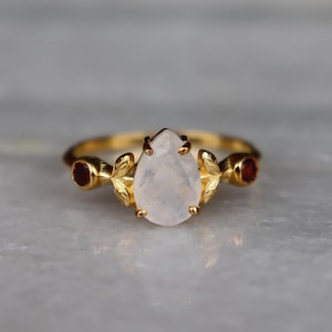 May include: A gold ring featuring a central pear-shaped, translucent white gemstone flanked by two small, round, deep red stones. The setting includes gold metal leaf-shaped accents on either side of the central stone, connected to a thin gold band.
