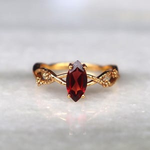 Könnte beinhalten: Ein goldener Ring mit einem mittig gefassten, tiefroten Granat im Marquise-Schliff. Die Ringschiene hat ein gedrehtes, rankenartiges Design mit kleinen, geprägten floralen Details auf der Metalloberfläche, die für eine strukturierte Optik sorgen.
