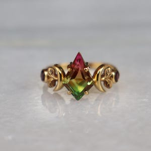 May include: A gold ring featuring a kite-shaped, multi-colored gemstone that transitions from pink at the top to green at the bottom. The band is decorated with crescent moon motifs on each side, accented by small, dark red round stones.