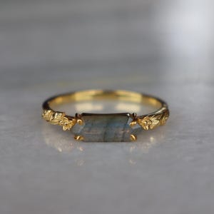 May include: A gold ring featuring a horizontal, rectangular, grey-blue labradorite gemstone set in a prong setting. The band is crafted with a detailed, raised leaf and floral pattern that extends along the sides of the central stone setting.
