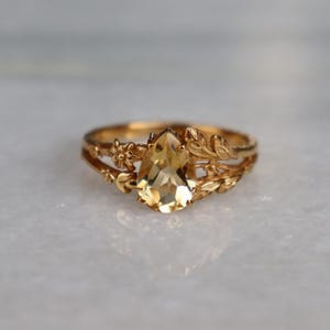 Può includere: Un anello in oro con una pietra preziosa gialla sfaccettata a forma di pera al centro. La fascia è decorata con dettagli metallici in rilievo che ricordano piccoli fiori e foglie che si estendono dalla pietra centrale lungo i lati dell'anello.