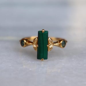 May include: A gold ring featuring a central rectangular green malachite stone set vertically. The band is adorned with small, sculpted leaf motifs on either side of the stone, leading to two smaller, round, dark green accent stones on the sides.