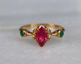 May include: A gold ring featuring a central marquise-cut red gemstone flanked by two small round green gemstones. The metal band is detailed with leaf-like motifs surrounding the central stone, creating a botanical appearance on the setting.