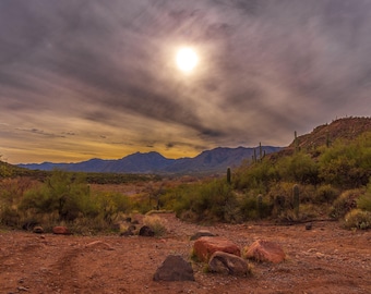 May include: A scenic view of a desert landscape with mountains in the distance. The sun is shining brightly through a cloudy sky. There are rocks and cacti in the foreground.