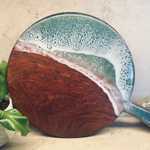May include: A round wooden cutting board with a resin pour that resembles an ocean wave. The wave is white with a teal blue base and a brown wooden base.