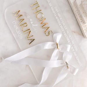 May include: A rectangular frosted acrylic wedding ring holder featuring gold lettering that reads THOMAS and MARINA, the date 17.05.2026, and the text THEREFORE WHAT GOD HAS JOINED TOGETHER, LET NOT MAN SEPARATE. Two white satin ribbons are attached.