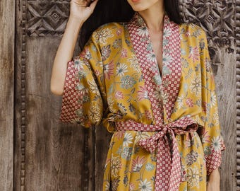 May include: A mustard yellow robe featuring a floral pattern in shades of white, pink, and green. The garment has a contrasting red and white geometric print on the lapels, cuffs, and a matching tie belt at the waist with a side slit.