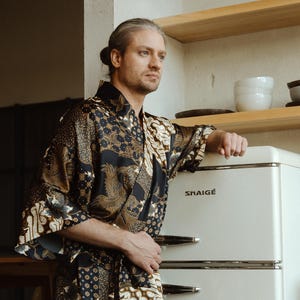 May include: A person wearing a black and brown patterned robe with a matching belt. The robe has a floral design and is open in the front. The person is standing next to a white refrigerator with the brand name "SHAIGE" on it.
