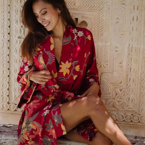 Könnte beinhalten: Ein roter Bademantel mit Blumenmuster und gelben und weißen Akzenten. Der Bademantel hat ein Wickeldesign und ist aus einem seidigen Material gefertigt. Das Blumenmuster umfasst stilisierte Blumen und Blätter. Der Bademantel wird offen getragen und zeigt ein schwarzes Oberteil.