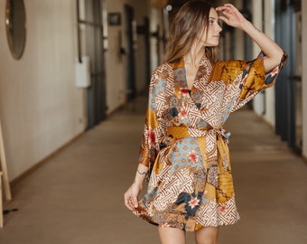 May include: A short kimono-style robe featuring a patchwork pattern of mustard yellow, light blue, and dark brown geometric shapes, floral motifs, and intricate line designs. The garment includes wide sleeves and a matching fabric tie belt.
