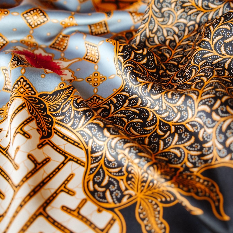 May include: A silk fabric featuring a detailed batik pattern with a light blue background, intricate black and gold floral motifs, and small red accents. The material is draped to show the texture and the repeating geometric and organic designs.
