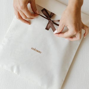 May include: A square off-white fabric envelope pouch featuring a brown ribbon tie closure at the top center. The word "m&egrave;nuo" is printed in brown lowercase letters on the front surface of the fabric pouch.