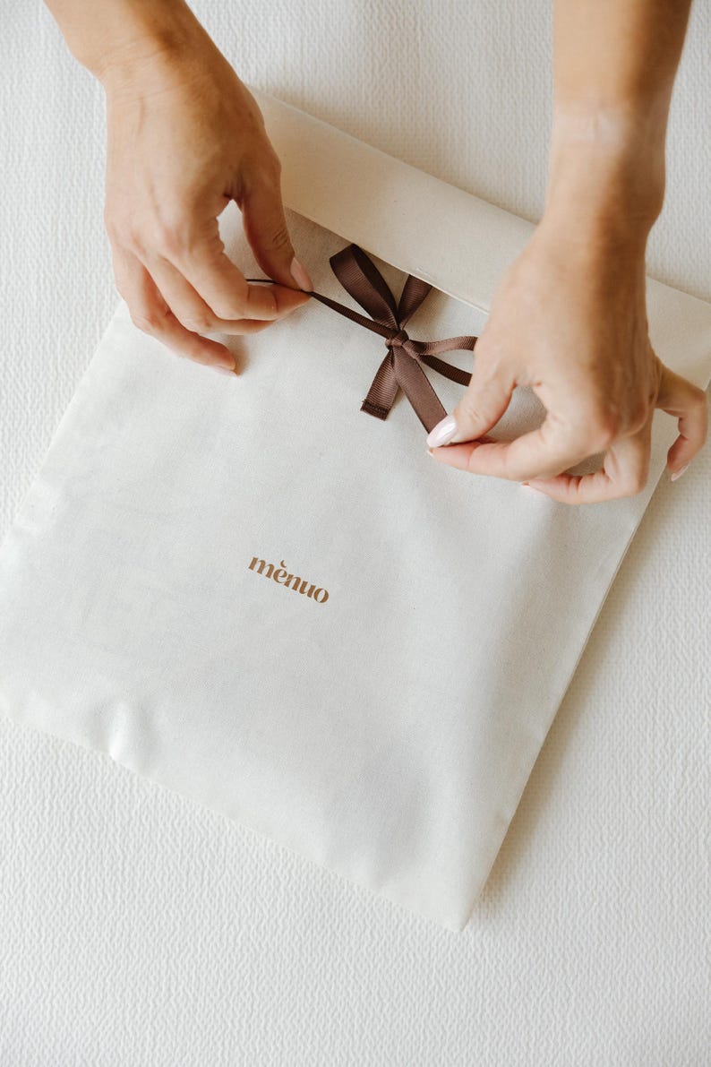 May include: A square off-white fabric envelope pouch featuring a brown ribbon tie closure at the top center. The word "m&egrave;nuo" is printed in brown lowercase letters on the front surface of the fabric pouch.
