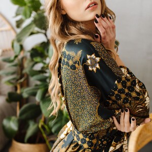 May include: A woman with long blonde hair wears a dark green and gold patterned silk robe. The robe has a floral design and a sash tied at the waist.