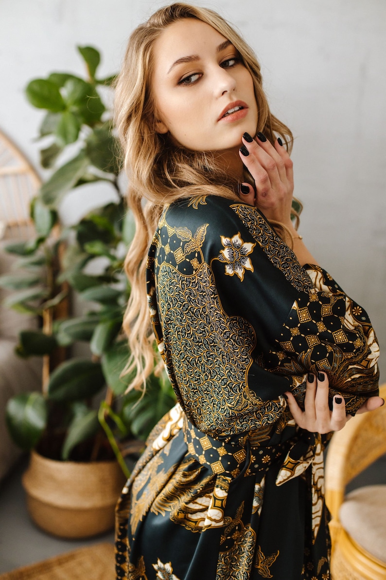 May include: A woman with long blonde hair wears a dark green and gold patterned silk robe. The robe has a floral design and a sash tied at the waist.
