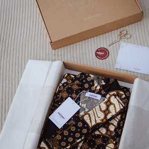 May include: A black and gold patterned fabric set with two pieces, folded and packaged in a brown box with the word "mimio" printed on the box. The fabric has a traditional Indonesian batik design.