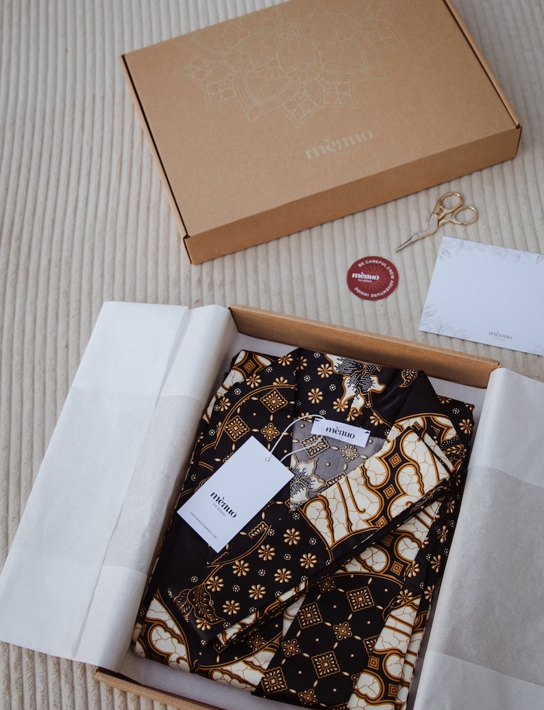 May include: A black and gold patterned fabric set with two pieces, folded and packaged in a brown box with the word "mimio" printed on the box. The fabric has a traditional Indonesian batik design.