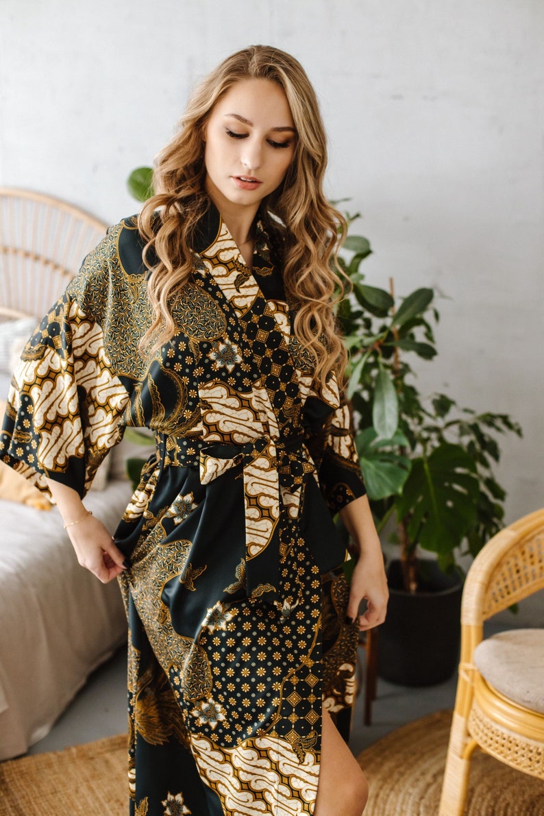 May include: A woman wearing a long, dark green and gold patterned robe with a sash tied at the waist. The robe has a traditional Indonesian batik design.