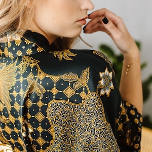 May include: A woman wearing a dark green and gold patterned silk robe. The robe has a traditional Indonesian batik design.