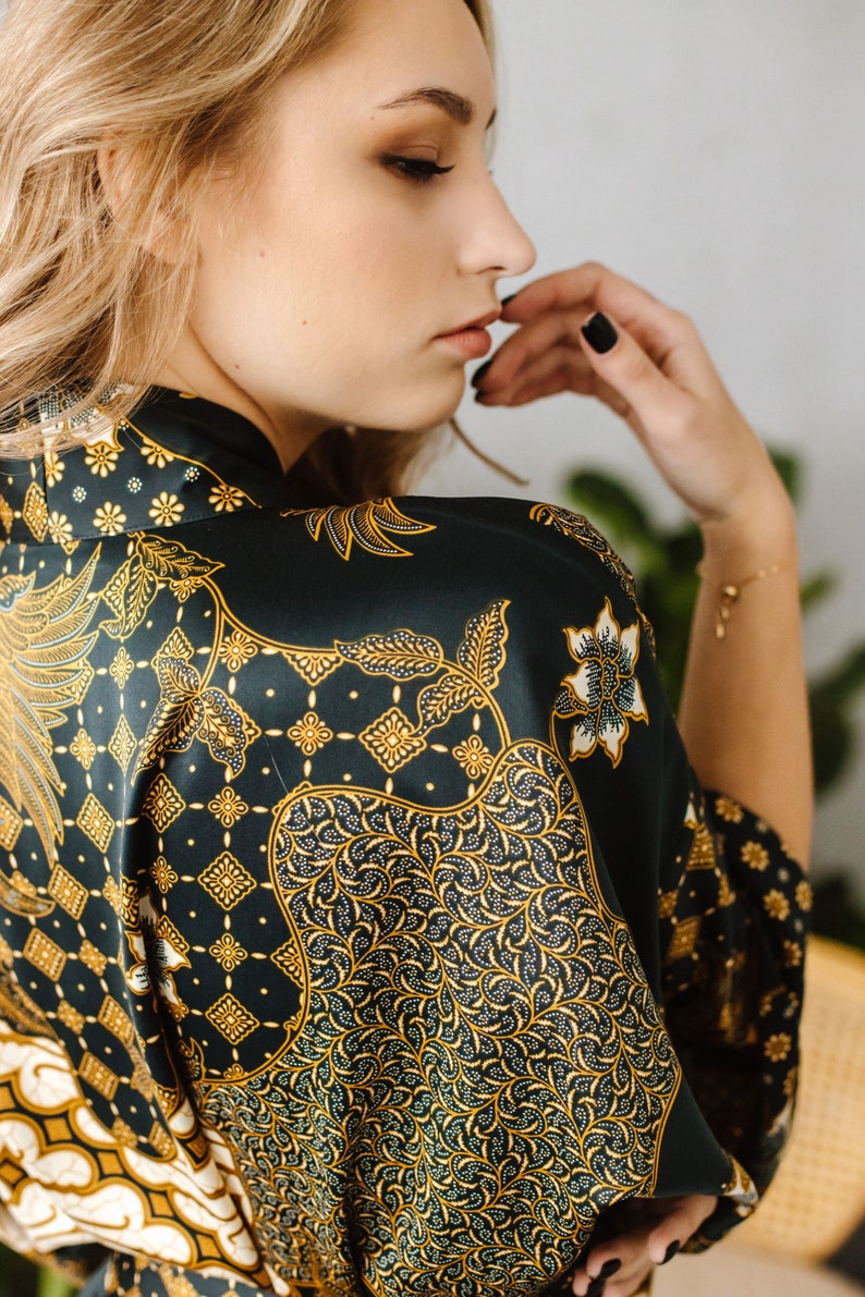 May include: A woman wearing a dark green and gold patterned silk robe. The robe has a traditional Indonesian batik design.