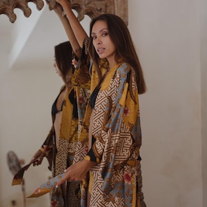 May include: A long-sleeved kimono robe featuring a vibrant mustard yellow, light blue, and black batik-inspired pattern with intricate geometric shapes and floral motifs. The garment has an open front design with wide sleeves and a relaxed fit.