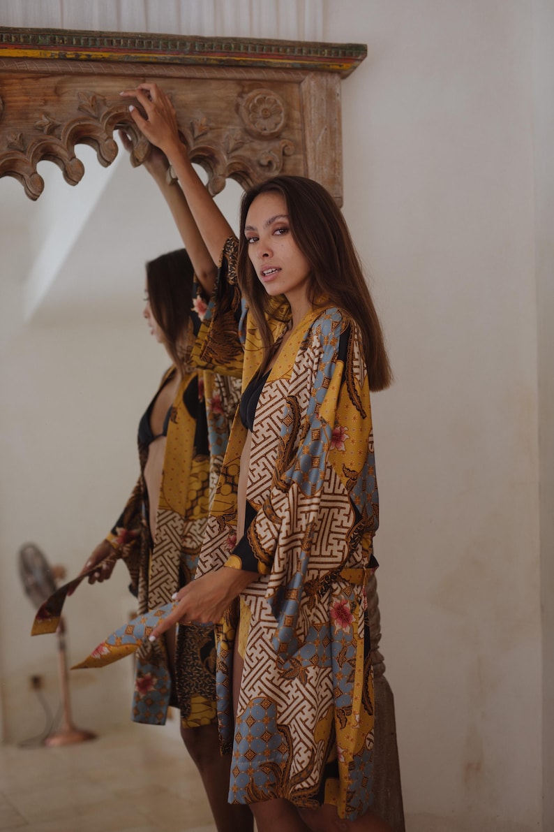 May include: A long-sleeved kimono robe featuring a vibrant mustard yellow, light blue, and black batik-inspired pattern with intricate geometric shapes and floral motifs. The garment has an open front design with wide sleeves and a relaxed fit.