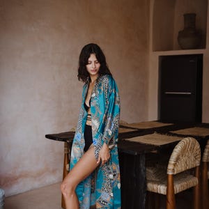 Puede incluir: Una mujer lleva una bata de seda azul turquesa con un estampado floral y geom&eacute;trico. La bata est&aacute; abierta, dejando ver un sujetador negro debajo. La mujer est&aacute; de pie en una habitaci&oacute;n con una mesa de madera y sillas.