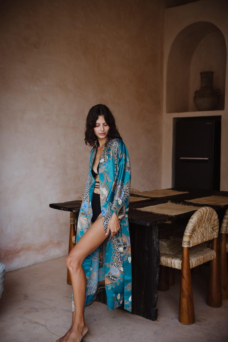 Puede incluir: Una mujer lleva una bata de seda azul turquesa con un estampado floral y geom&eacute;trico. La bata est&aacute; abierta, dejando ver un sujetador negro debajo. La mujer est&aacute; de pie en una habitaci&oacute;n con una mesa de madera y sillas.