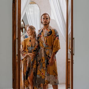 May include: Two matching mustard yellow robes featuring a patchwork design with floral motifs, geometric patterns, and dark blue accents. The robes have long sleeves, a wrap-front style with a tie belt, and reach approximately to the knee.