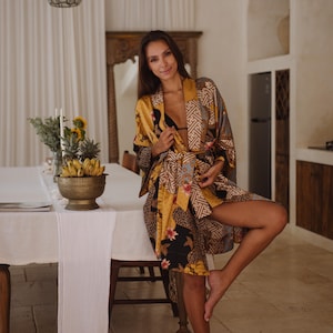 May include: A kimono-style robe featuring a vibrant mix of mustard yellow, black, and blue panels with intricate floral and geometric patterns. The garment includes wide sleeves, a matching fabric tie belt, and a wrap-front design.