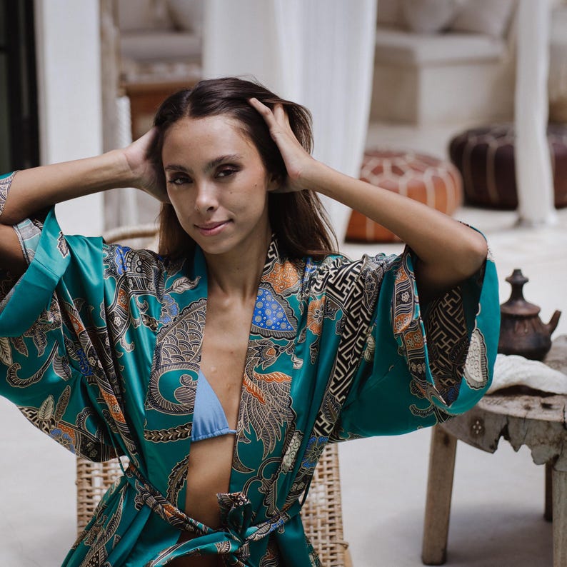 Puede incluir: Una mujer lleva una bata de seda azul turquesa con un estampado floral y geom&eacute;trico. La bata tiene un cintur&oacute;n para atar a la cintura.