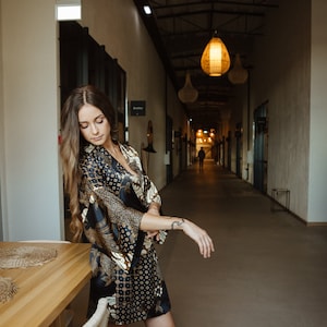 May include: A woman wearing a black and gold patterned silk robe stands in a hallway with a wooden table and chairs in the foreground. The hallway has a high ceiling with exposed beams and several hanging light fixtures.