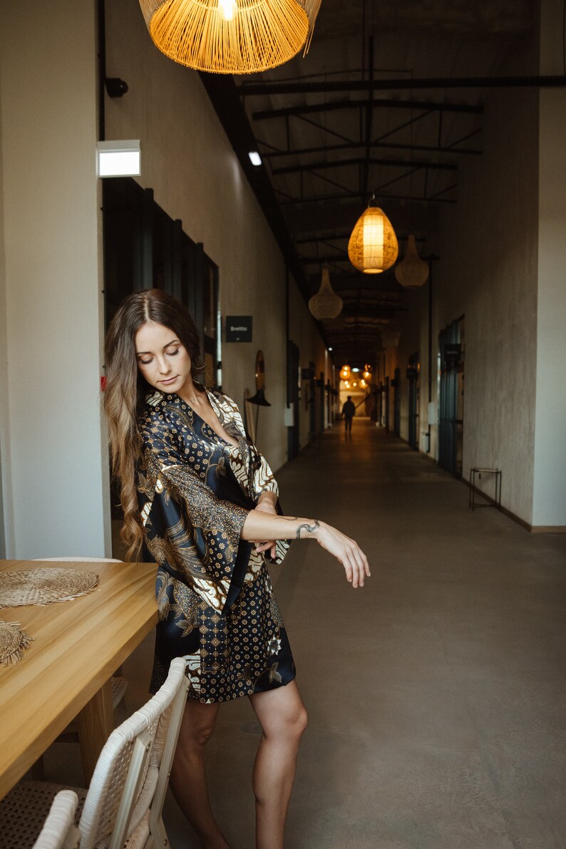 May include: A woman wearing a black and gold patterned silk robe stands in a hallway with a wooden table and chairs in the foreground. The hallway has a high ceiling with exposed beams and several hanging light fixtures.