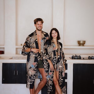 May include: A couple wearing matching black and gold patterned robes with intricate designs. The robes have a relaxed fit and are open in the front. The couple is standing in a kitchen with white walls and a black countertop.