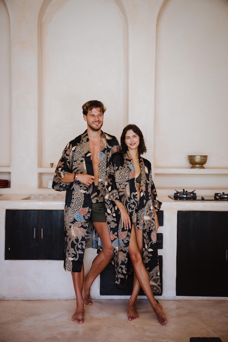 May include: A couple wearing matching black and gold patterned robes with intricate designs. The robes have a relaxed fit and are open in the front. The couple is standing in a kitchen with white walls and a black countertop.