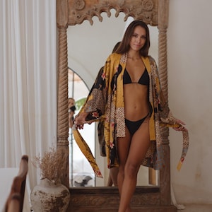 May include: A long-sleeved kimono robe featuring a patchwork design with mustard yellow, black, and blue panels. The fabric displays a mix of floral patterns, geometric shapes, and intricate motifs. It includes a matching tie belt at the waist.