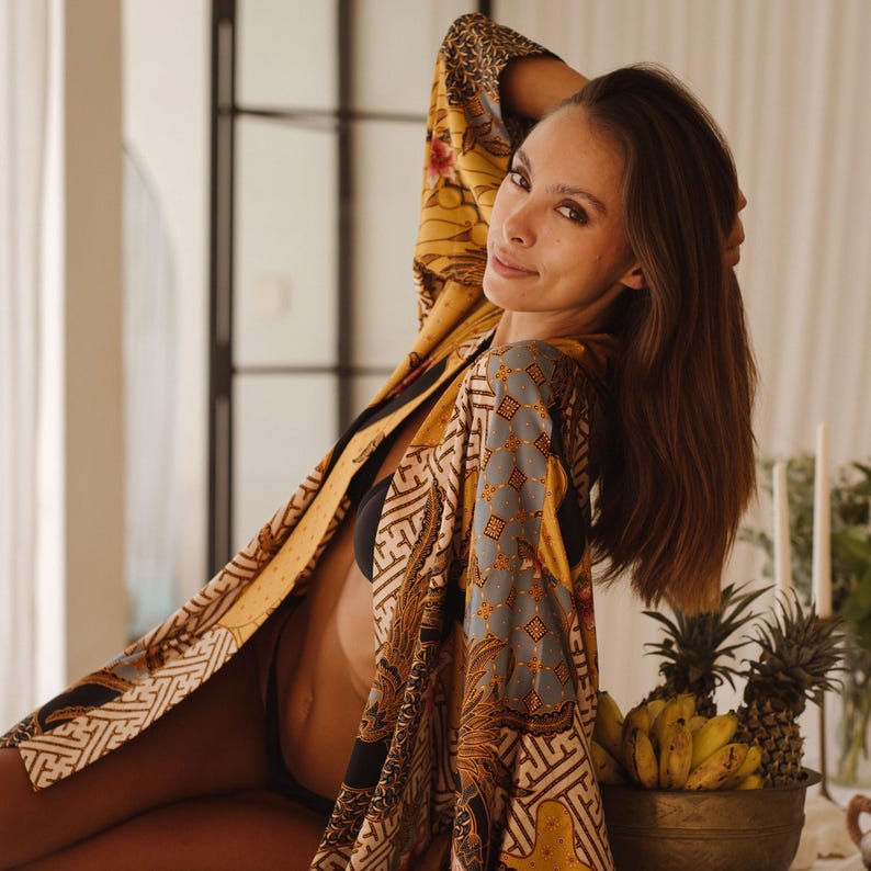 May include: A long-sleeved robe featuring a vibrant mustard yellow, blue, and black batik-inspired pattern with intricate geometric and floral motifs. The garment is worn open over a black undergarment, showcasing a flowing, lightweight fabric.