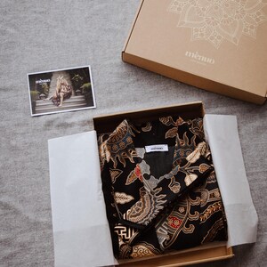 May include: A black and gold patterned fabric shirt with a white label that says "memo." The shirt is folded and placed in a brown cardboard box with a white mandala design and the word "memo."
