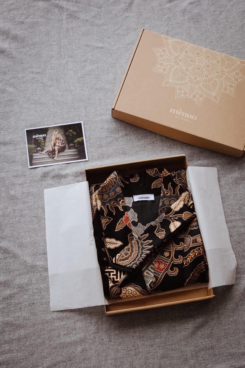 May include: A black and gold patterned fabric shirt with a white label that says "memo." The shirt is folded and placed in a brown cardboard box with a white mandala design and the word "memo."