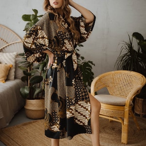 May include: A woman wearing a long black and gold patterned robe with a sash. The robe has a side slit and is made of a silky material. The woman is standing on a woven rug in front of a wicker chair.