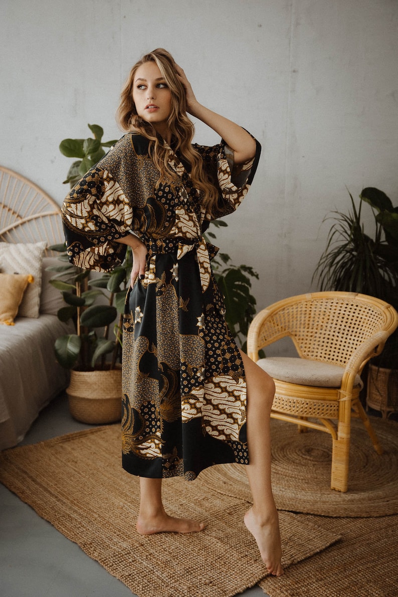May include: A woman wearing a long black and gold patterned robe with a sash. The robe has a side slit and is made of a silky material. The woman is standing on a woven rug in front of a wicker chair.