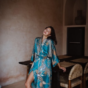 Puede incluir: Una mujer lleva una bata de seda azul turquesa con estampado floral. La bata tiene un cintur&oacute;n de lazo y mangas largas. La mujer est&aacute; de pie en una habitaci&oacute;n con una mesa de madera y sillas.