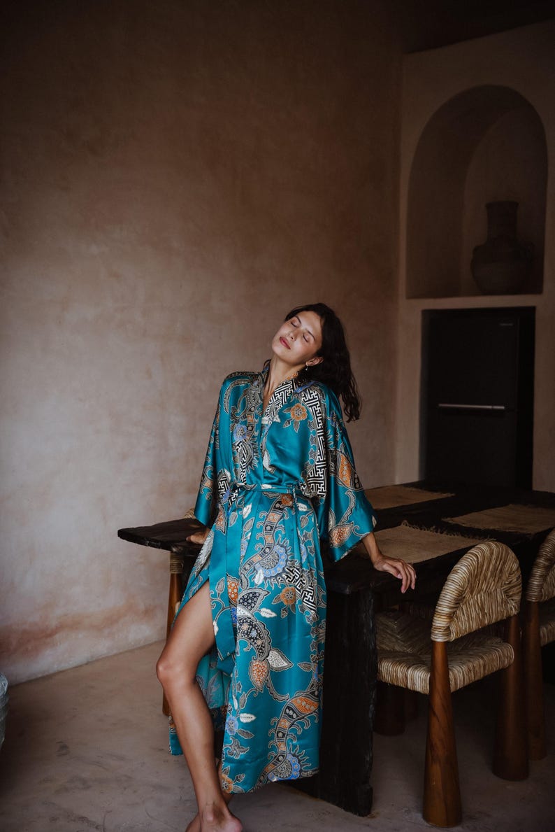 Puede incluir: Una mujer lleva una bata de seda azul turquesa con estampado floral. La bata tiene un cintur&oacute;n de lazo y mangas largas. La mujer est&aacute; de pie en una habitaci&oacute;n con una mesa de madera y sillas.