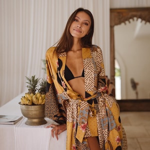 May include: A mustard yellow robe featuring a patchwork design of black, white, and blue geometric patterns mixed with floral motifs. The garment has long sleeves, a tie-waist belt, and a front opening worn over a black bikini top.