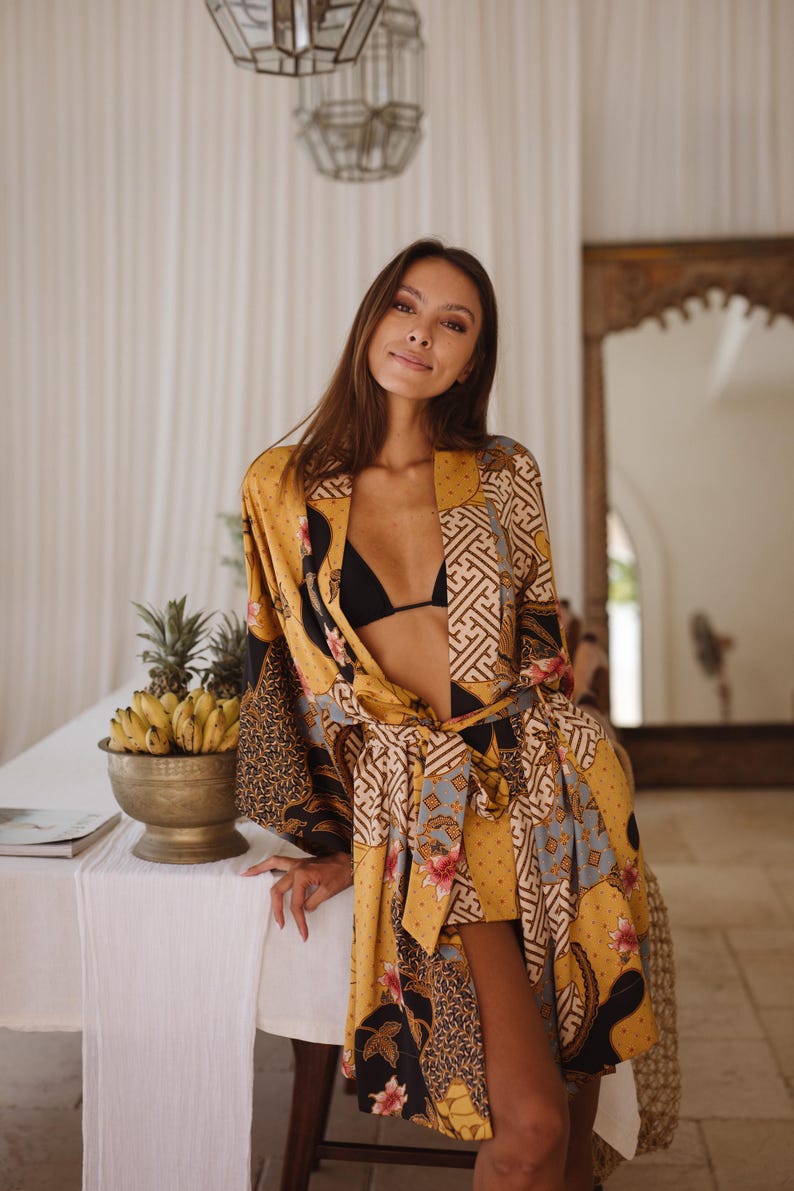 May include: A mustard yellow robe featuring a patchwork design of black, white, and blue geometric patterns mixed with floral motifs. The garment has long sleeves, a tie-waist belt, and a front opening worn over a black bikini top.