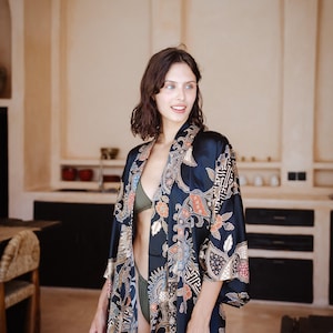 May include: A woman wearing a black silk robe with a colorful floral pattern. The robe has a shawl collar and long sleeves. The woman is standing in a room with a white wall and a wooden floor.